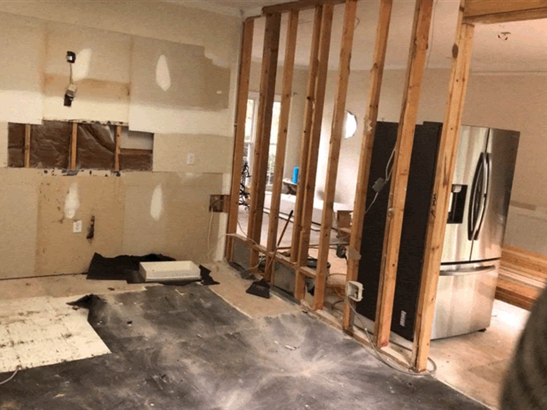 Interior view of a home renovation. Bare wood framing stands in place of walls, exposing wires, and a stainless steel refrigerator is visible.