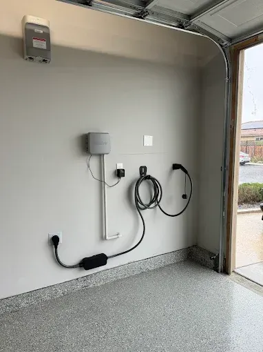 Electric vehicle charging equipment mounted on a neutral garage wall, featuring a coiled charging cable and power brick.