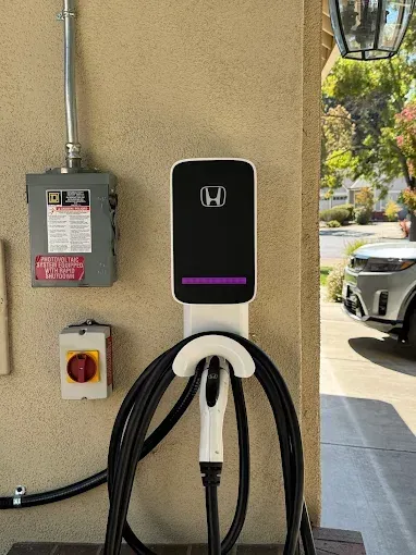 A Honda electric vehicle charger wall-mounted on a beige wall next to a grey electrical box and a red power switch.