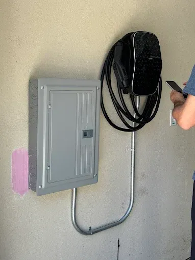 A gray electrical subpanel mounted next to a wall-mounted EV charger with a coiled black cable on an indoor wall.