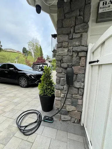 A black electric vehicle charger is mounted to a stone pillar, with its charging cable coiled on the paved driveway.
