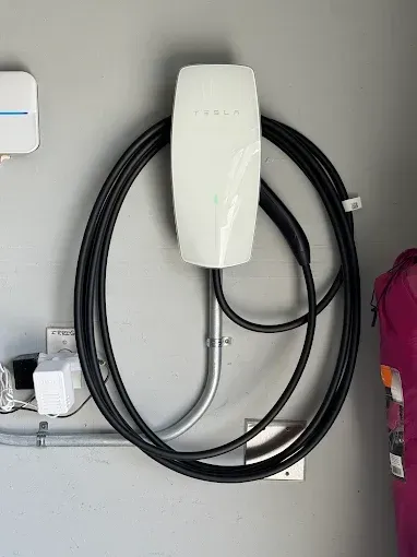 A white Tesla Wall Connector electric vehicle charger mounted on a wall with its black charging cable coiled around it.