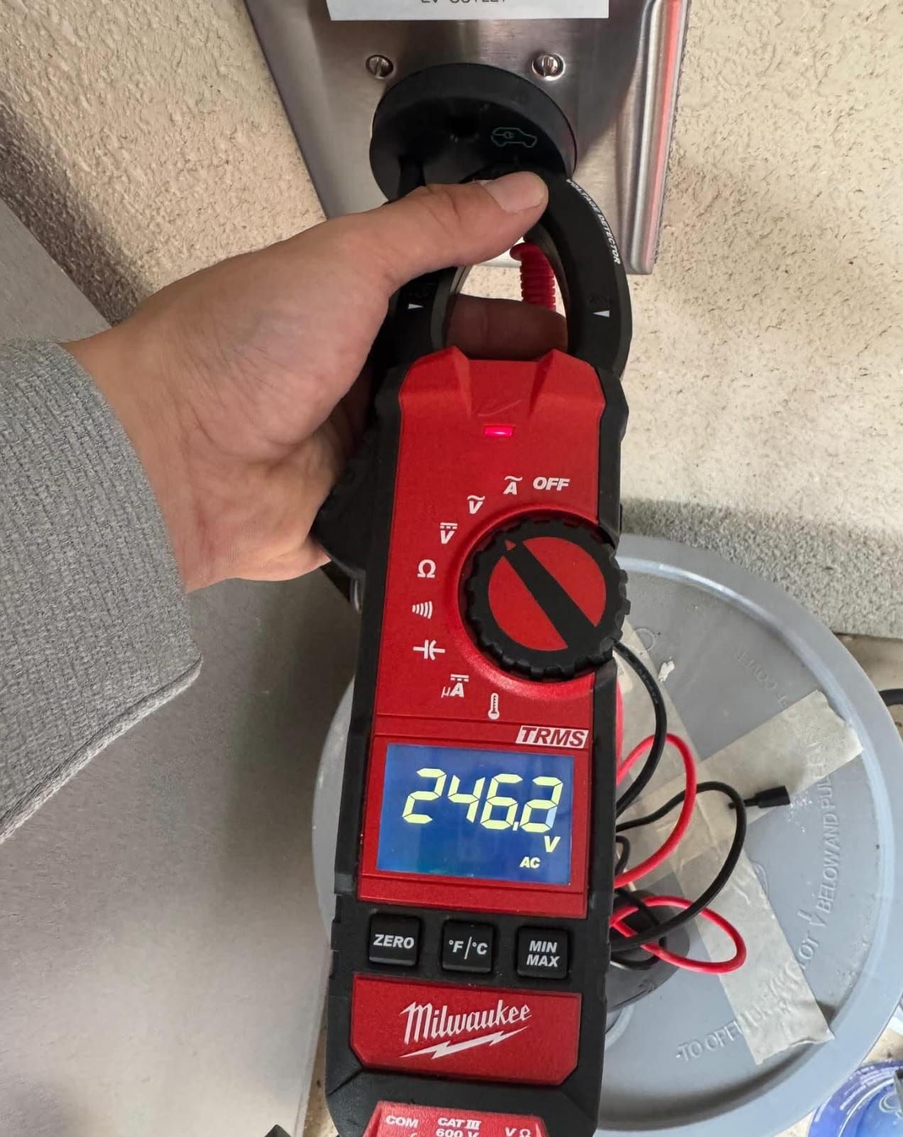 A hand holds a Milwaukee clamp meter measuring 246.2 V AC on an electrical connection.