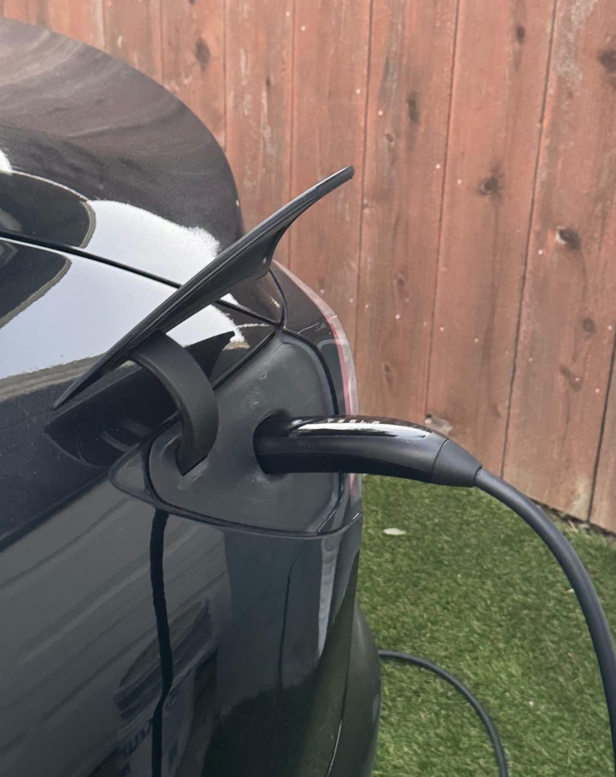 A close-up of a black electric vehicle being charged by a black charging cable, with a wooden fence in the background.