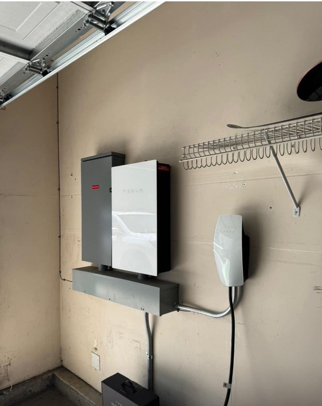 A Tesla Wall Connector and a home battery system mounted on a beige garage wall with a white wire rack above.