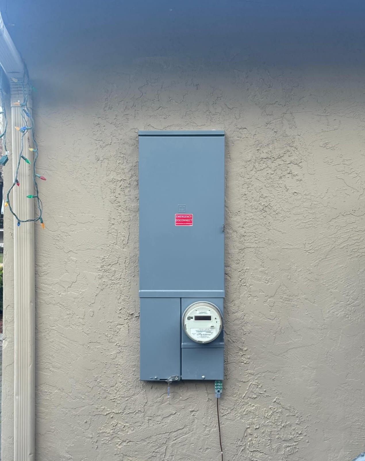 An electrical meter and service panel box mounted on a textured beige exterior wall.