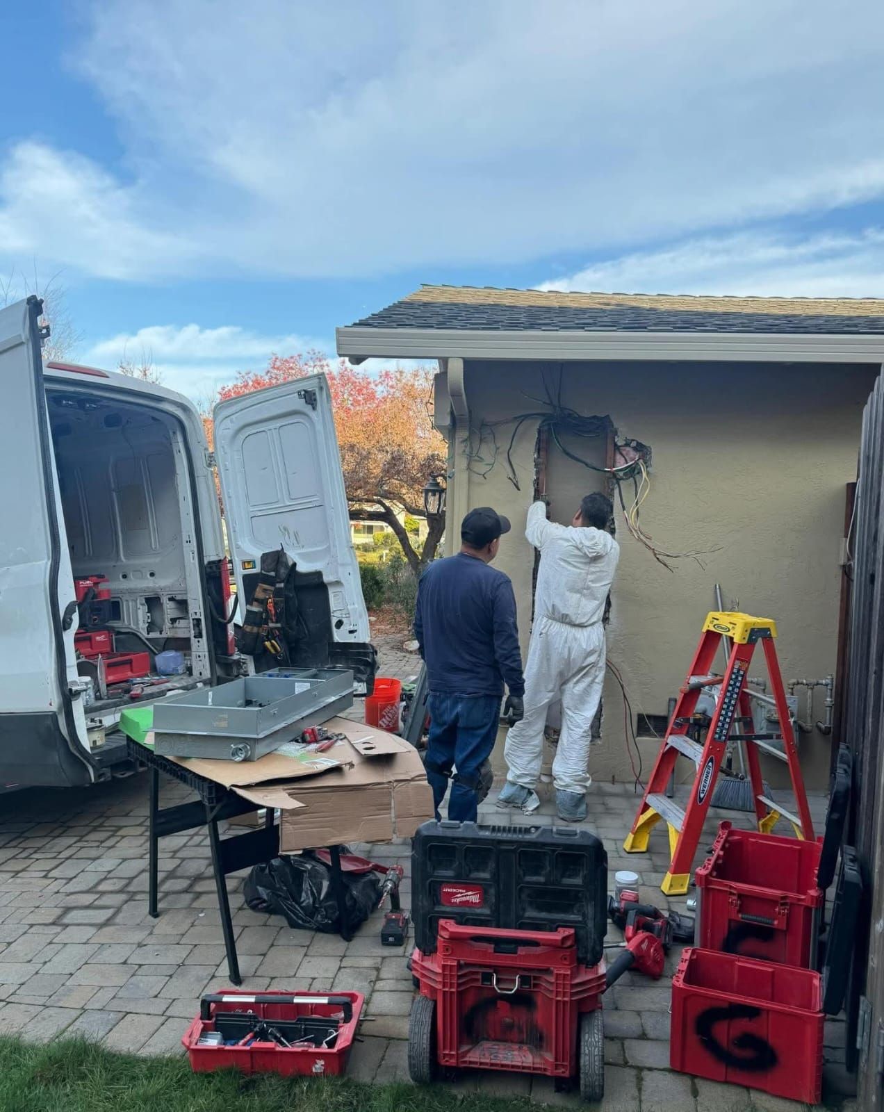 Two technicians in work gear perform repairs on an exterior wall near a service van, a folding table, and equipment cases.