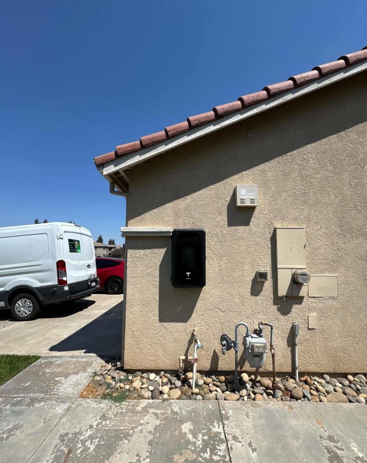 Exterior wall of a house with a black electric vehicle charger, utility pipes, a gas meter, and a van parked in the driveway.