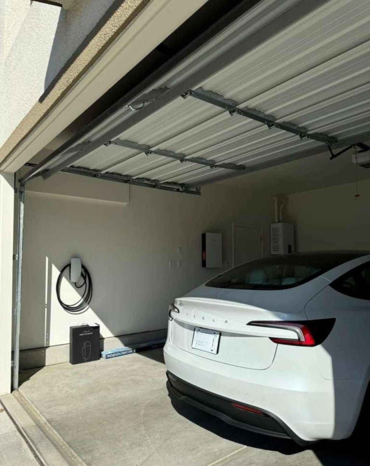 A white Tesla parked inside a garage, with a wall-mounted charging station and coiled cable visible to the left.