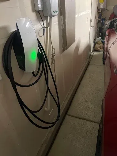 A Tesla Wall Connector mounted on a white garage wall with a green status light and coiled black charging cable.