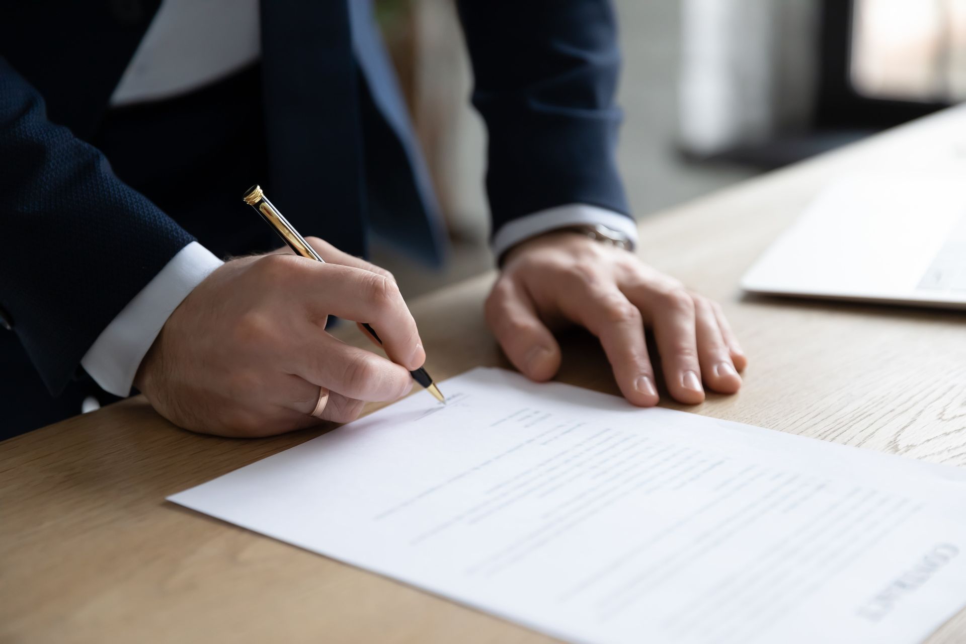 A person in a suit signing a document with a gold pen at a wooden desk.