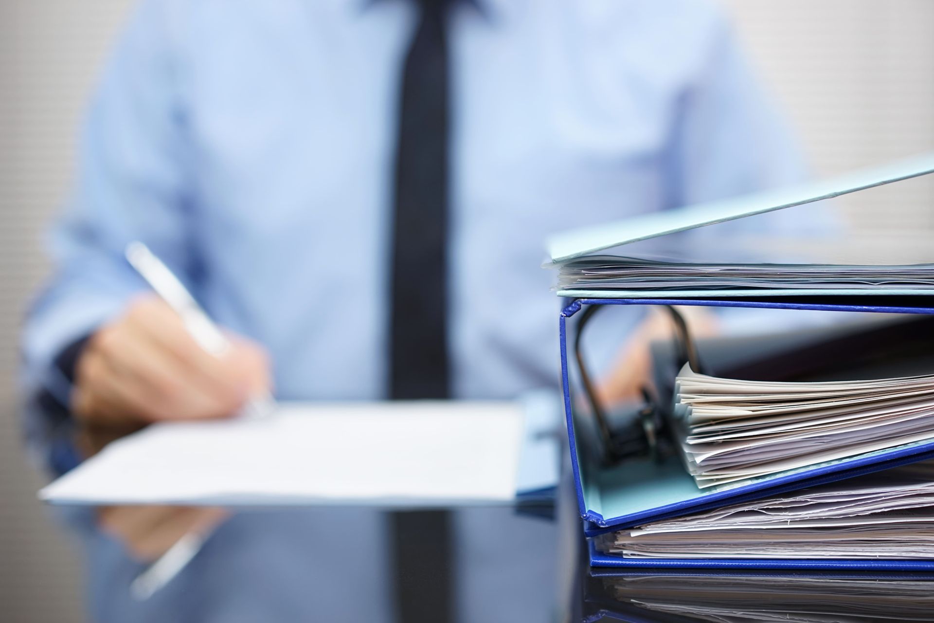 A person in a blue shirt and black tie writing at a desk, with a stack of blue binders in the foreground.