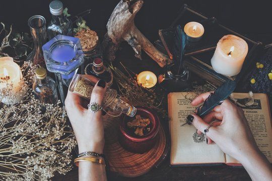 Hands pouring liquid from a bottle into a small bowl on a table with candles, potion bottles, and a book.
