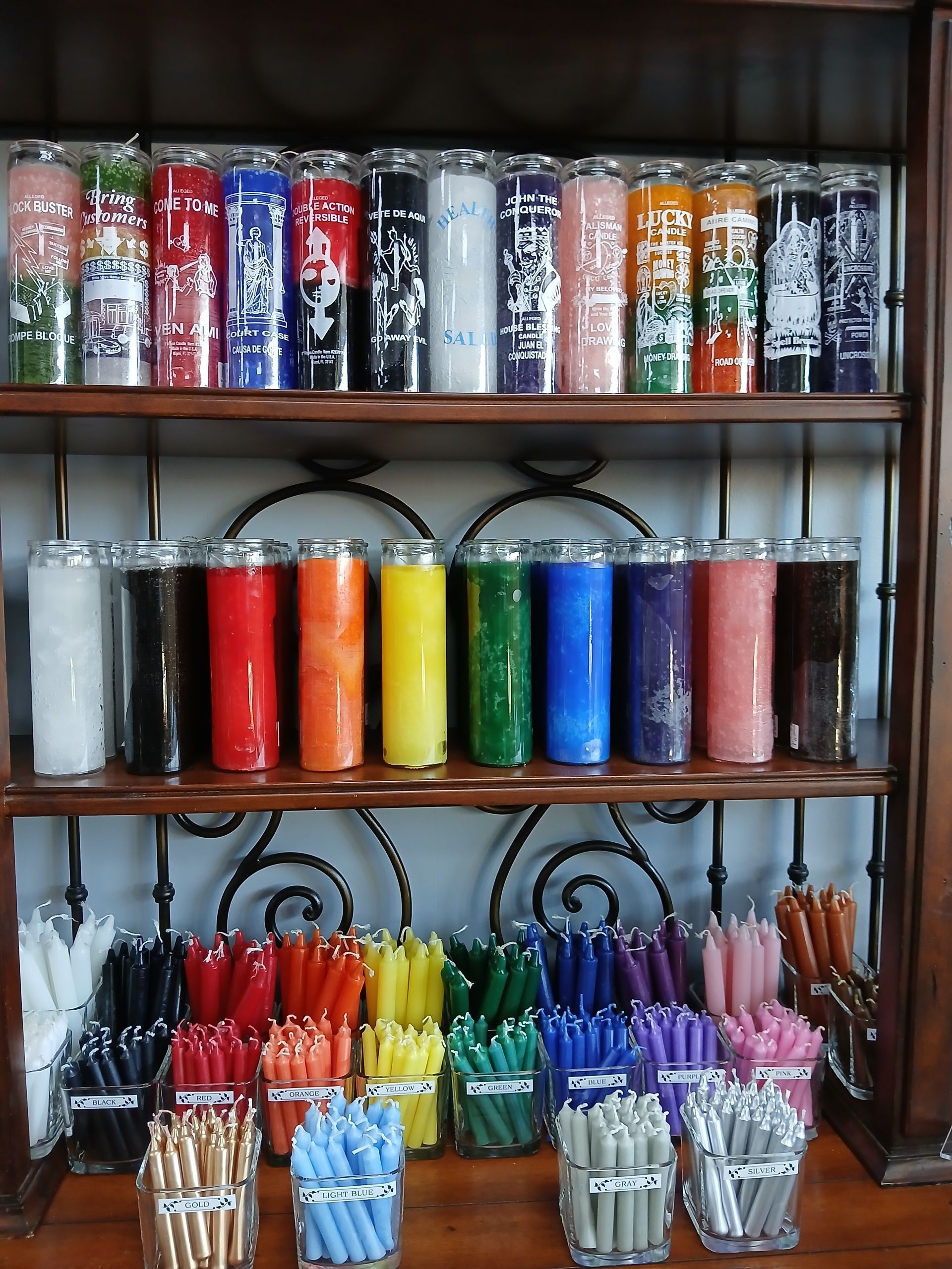 A wooden shelf displays various tall, colorful prayer candles and smaller taper candles.