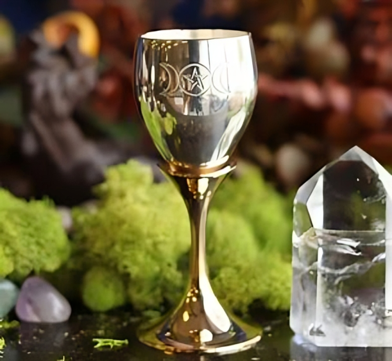 A golden chalice with the Triple Moon symbol is set amongst moss, crystals, and other 