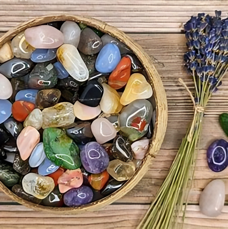 A wooden bowl filled with colorful polished gemstones next to a bundle of lavender and two loose stones on a wooden surface.