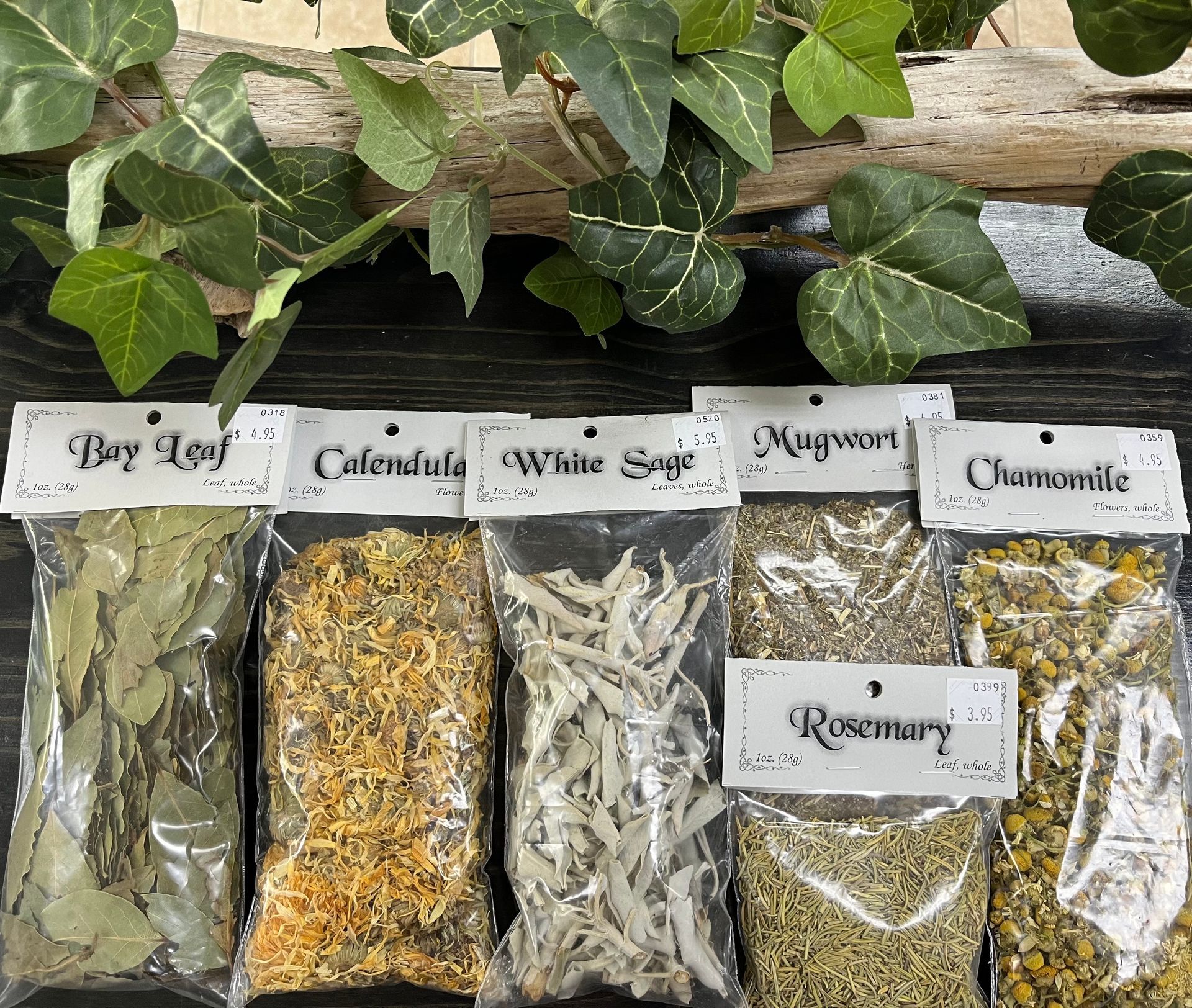 Bags of herbs: bay leaf, calendula, white sage, mugwort, chamomile, and rosemary, displayed on a wooden surface with ivy.