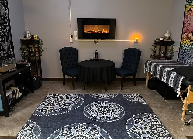 A dimly lit room with a round table and two chairs, rug, electric fireplace, two massage tables, and decorative shelves.