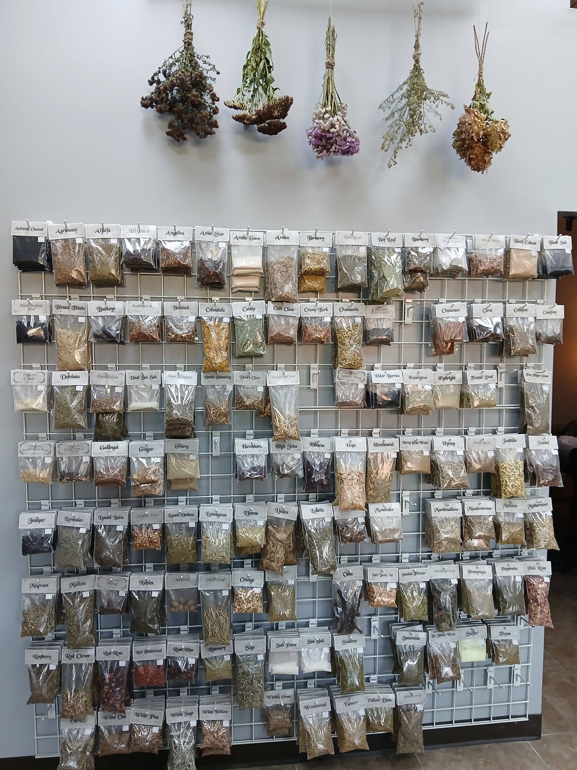 Three bottles of essential oil are sitting on a table next to flowers. Wall display of various herbs and seeds in clear bags, organized on a grid. Dried flower bundles hang above.