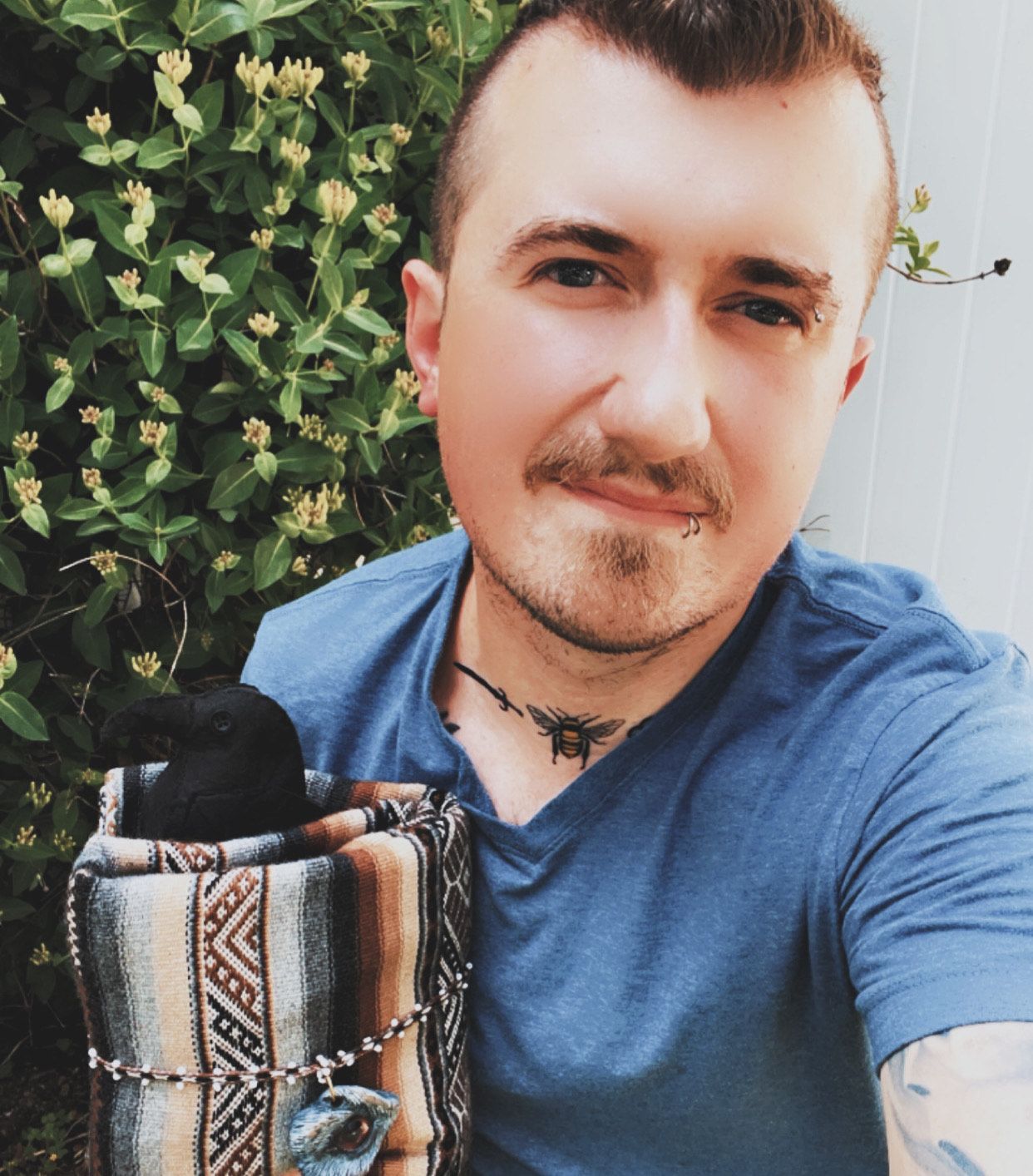 Man in blue shirt holding a patterned bag with a small black animal peeking out, set outside with green foliage.