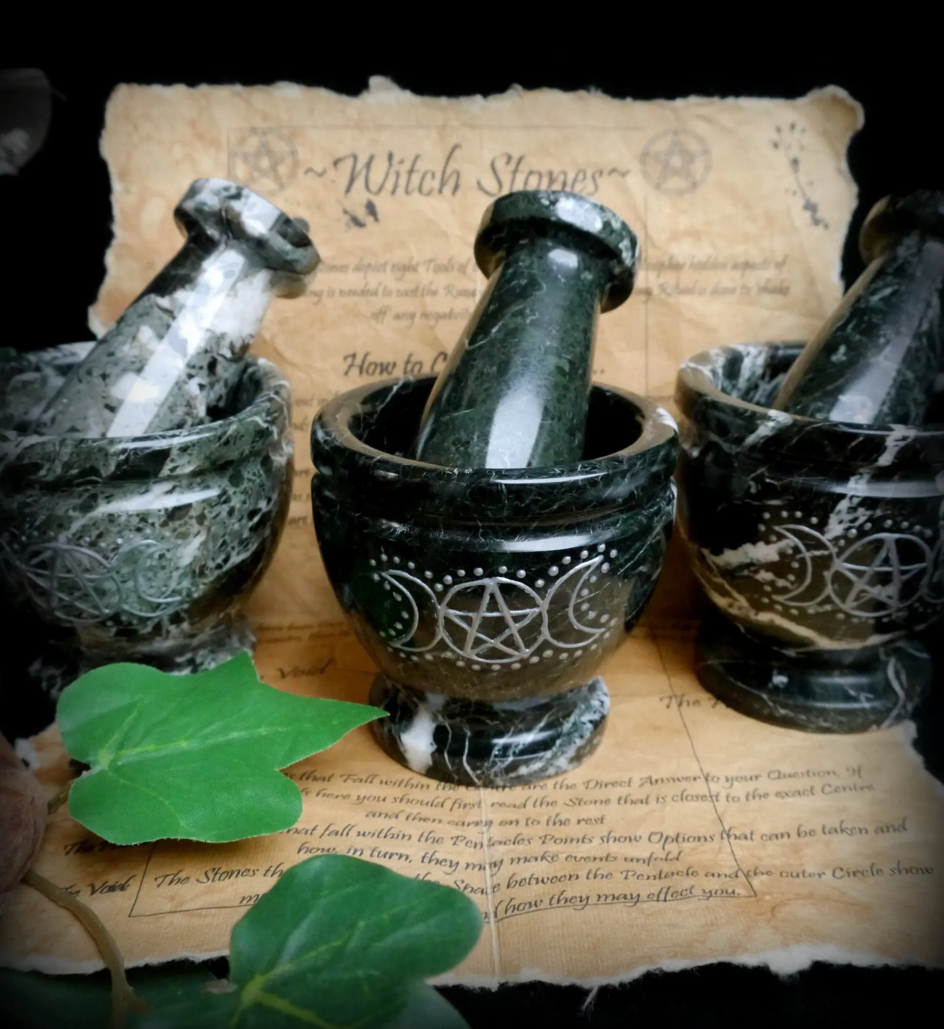 Three dark green mortar and pestle sets with etched symbols sit on aged parchment. Ivy leaf in foreground.