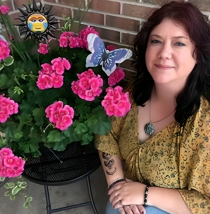 Woman sits by pink flowers, a sun decoration, and a butterfly. She wears a yellow shirt and has a tattoo on her arm.