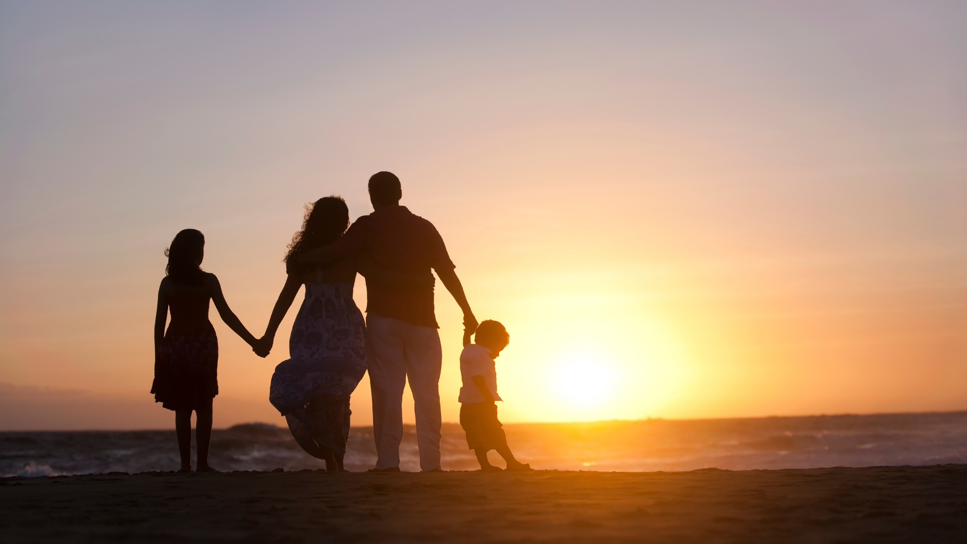 A family of four stands on a beach at sunset, silhouetted against the orange sky and ocean.