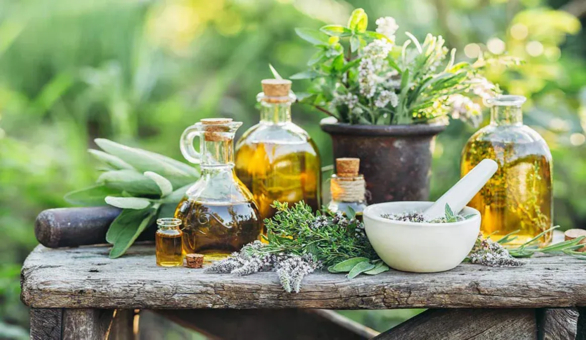 Bottles of oil with herbs, mortar and pestle, and plants on a weathered wooden surface.