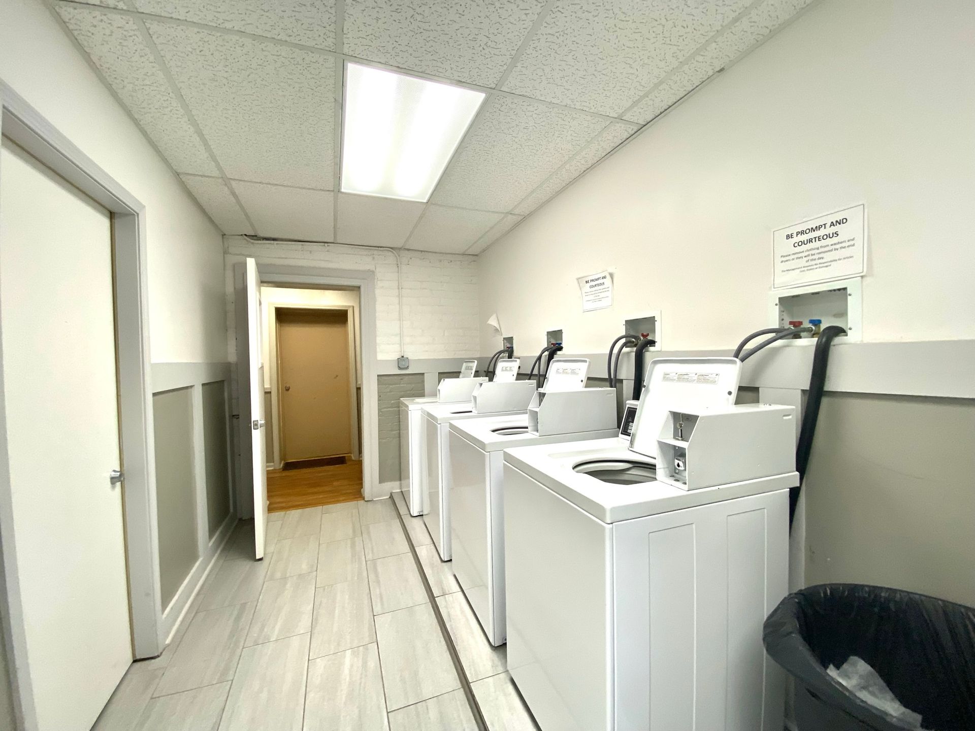 A laundry room with a row of washing machines and a trash can.