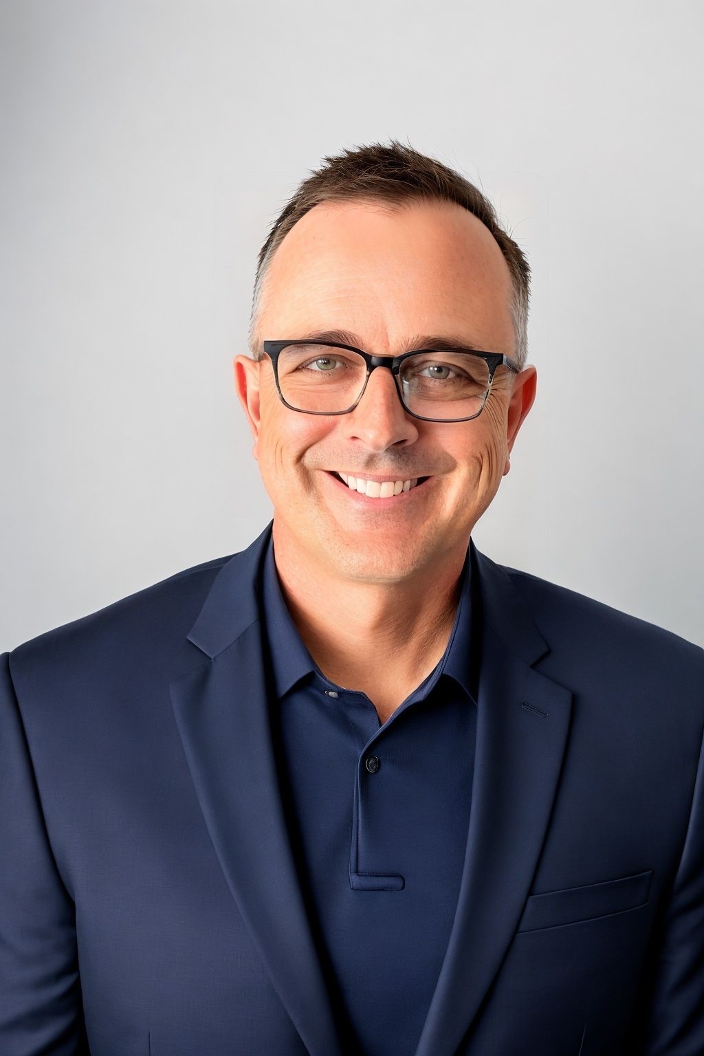 Man with glasses smiling, wearing a navy blue blazer and shirt, against a light gray backdrop.