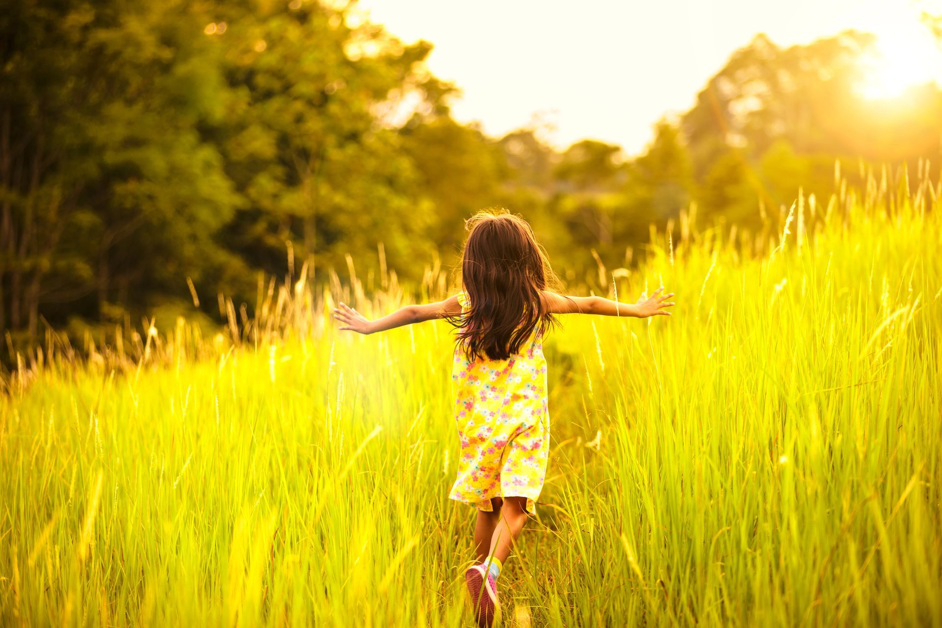 Girl with arms outstretched running through tall, sunlit grass.
