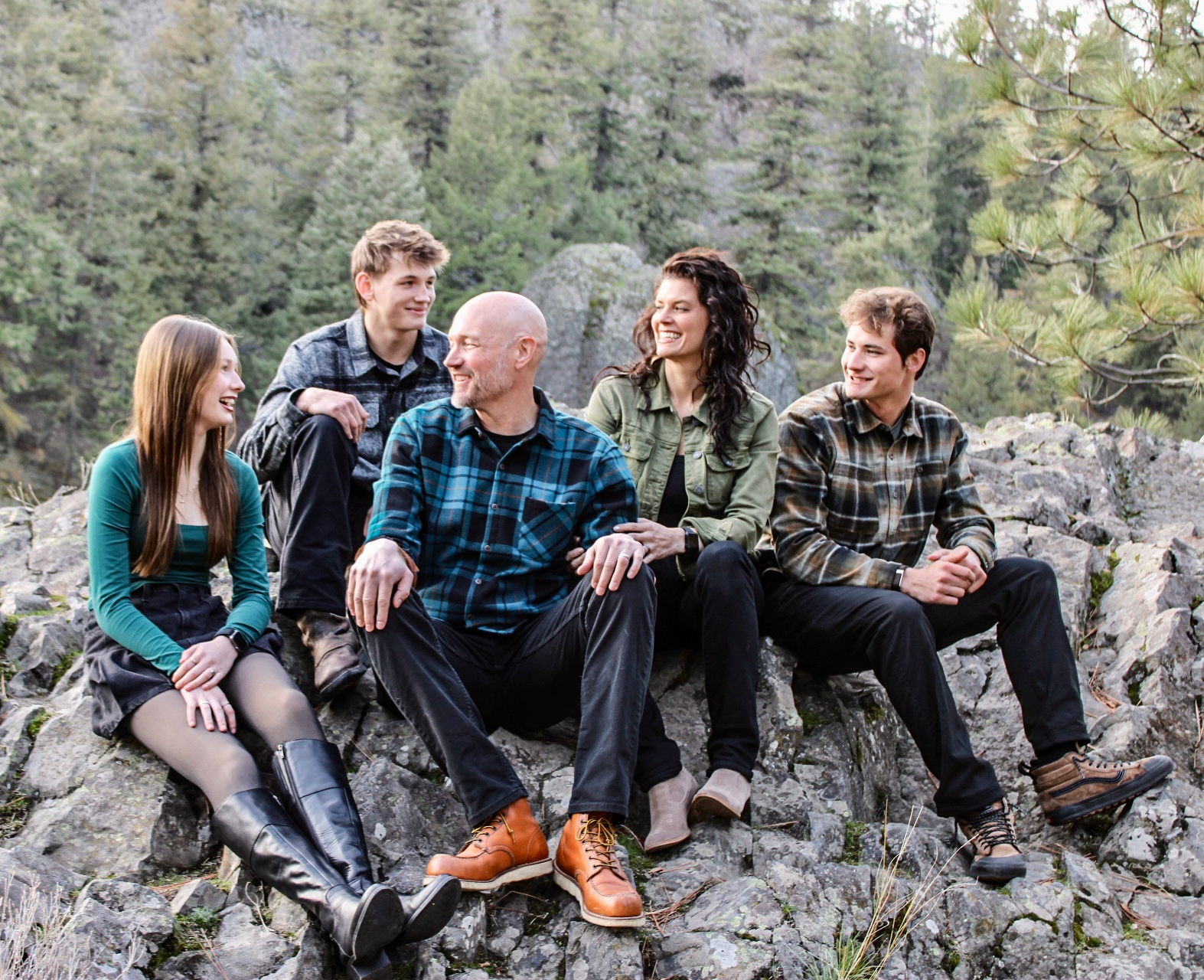 Family of five sitting on a rock outdoors; smiling, wearing casual clothes.