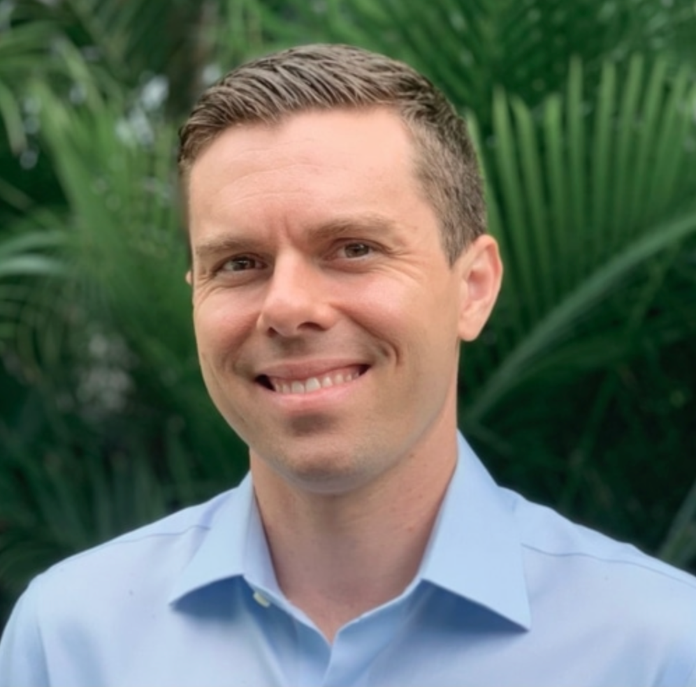Man in a light blue shirt smiles at the camera, with a blurred green background of foliage.
