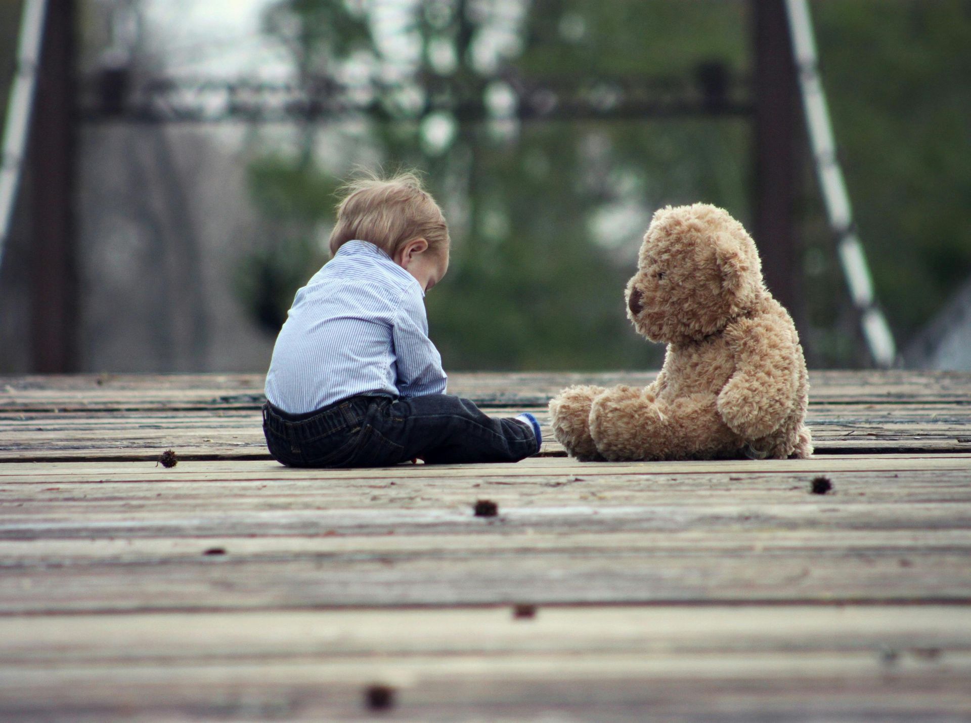 Child sitting on wooden bridge, facing a teddy bear. Blue shirt, jeans, overcast day.