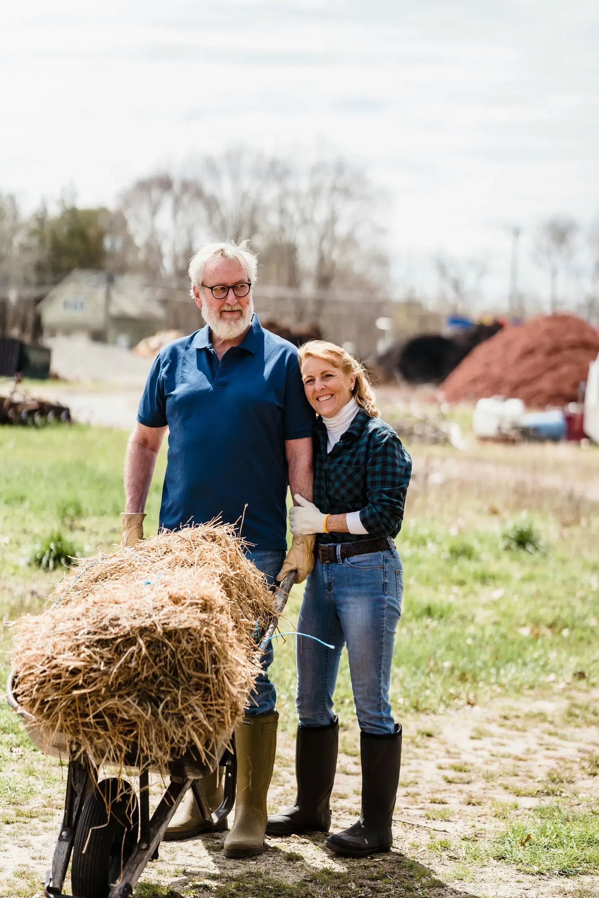 Couple poses with wheelbarrow of hay, outdoors. Man in blue shirt, woman in plaid, both in boots.