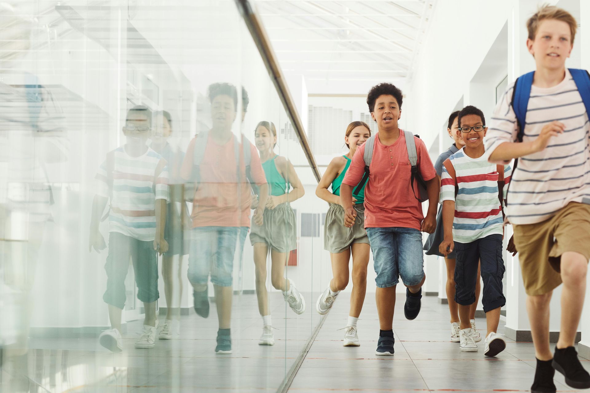 Children run down a school hallway; some blurred, wearing backpacks and casual clothes, with bright natural lighting.