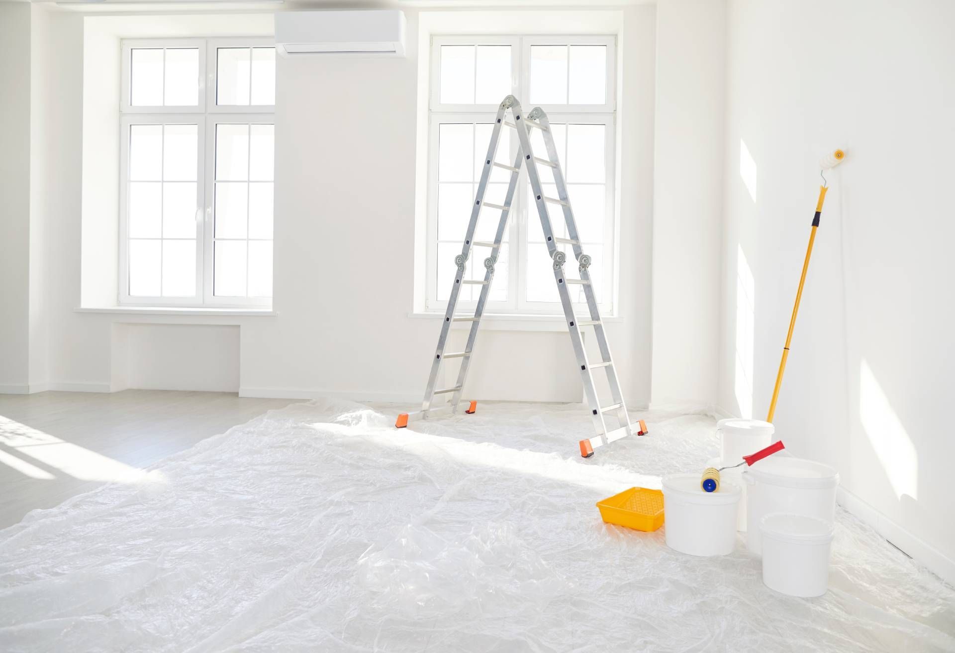 Bright, empty room with plastic-covered floor, ladder, paint supplies, and open windows during renovation