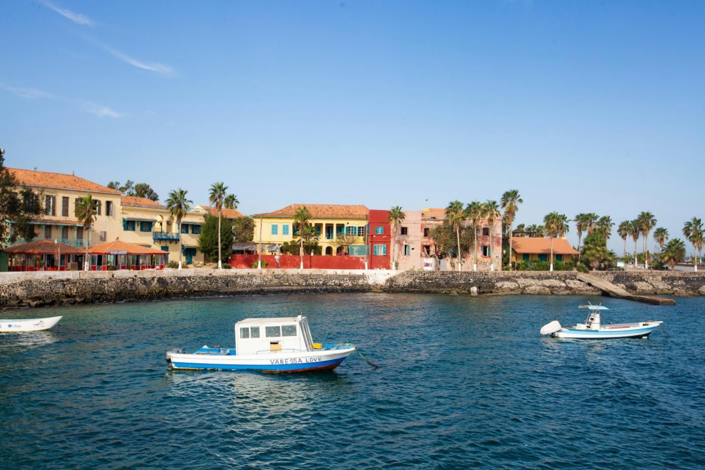 Boats on blue water in front of a colorful coastal town with palm trees and a clear blue sky.