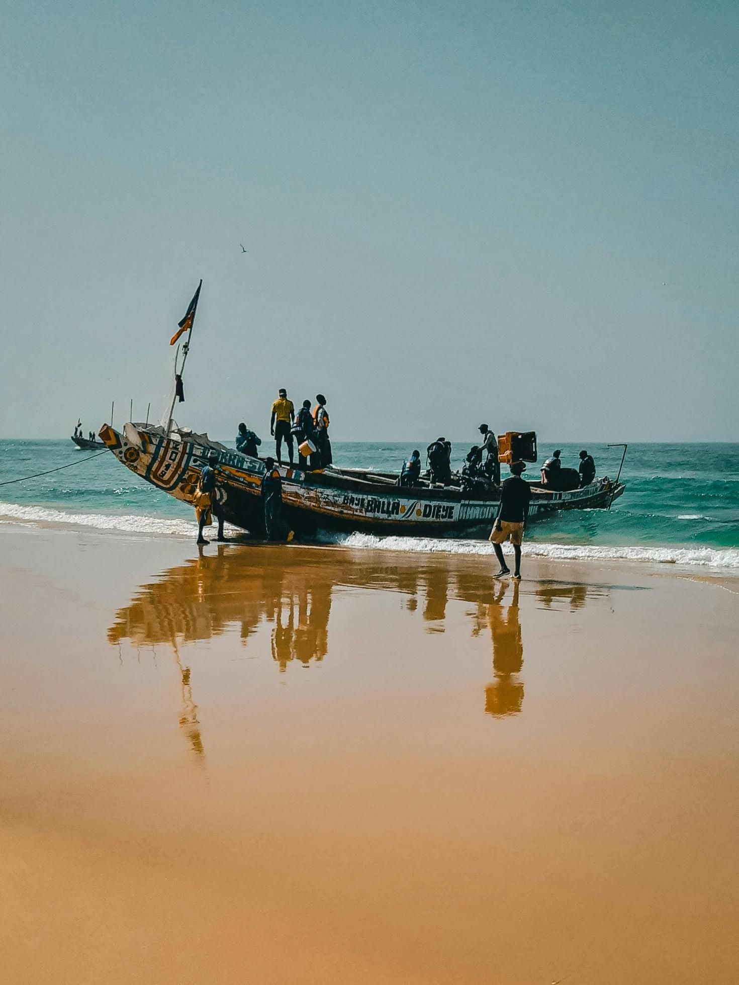 Boat with several people on a sandy beach. Boat's reflection visible in wet sand. Blue sky and ocean in background.
