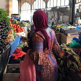 Woman in a red headscarf and patterned dress shops at a market stall filled with produce.