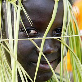 A person with very dark skin smiles, framed by woven green leaves.