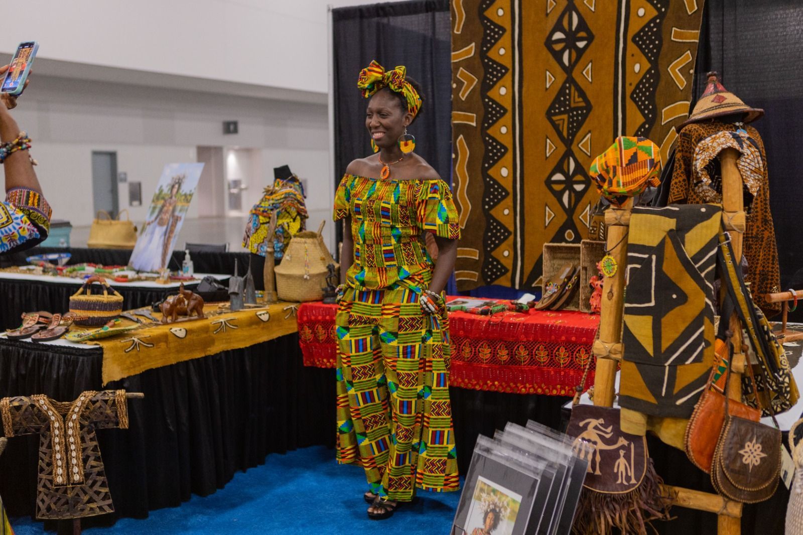 Woman in colorful African dress smiles, standing near a display of textiles and clothing.