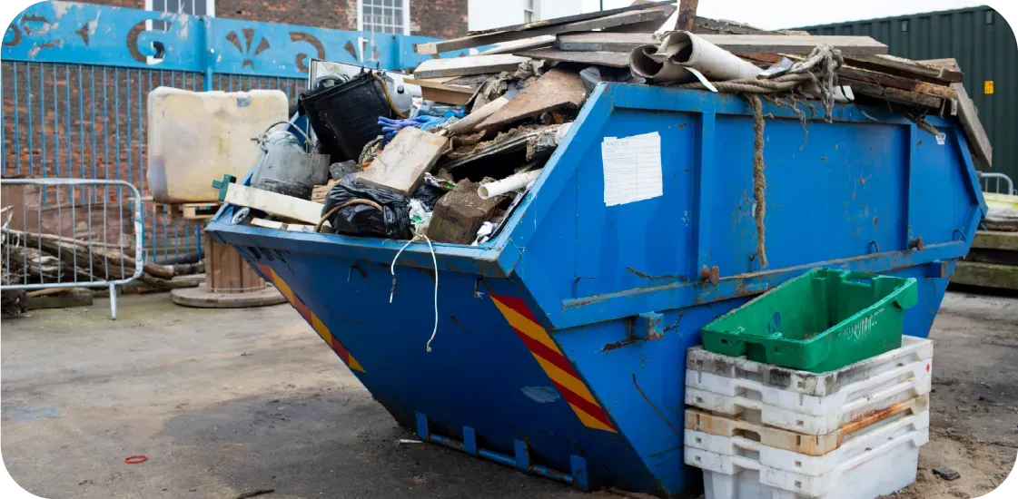 A blue dumpster overflowing with trash, outdoors. Green bins and white crates are beside it.