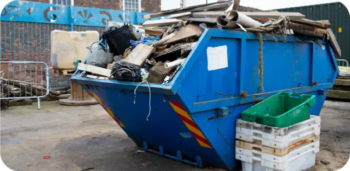 A blue dumpster overflowing with trash, outdoors. Green bins and white crates are beside it.