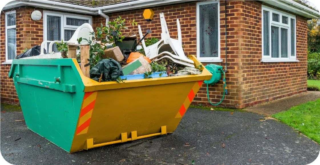 Green and yellow dumpster overflowing with trash, in front of a brick house.