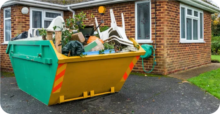 Green and yellow dumpster overflowing with trash, in front of a brick house.
