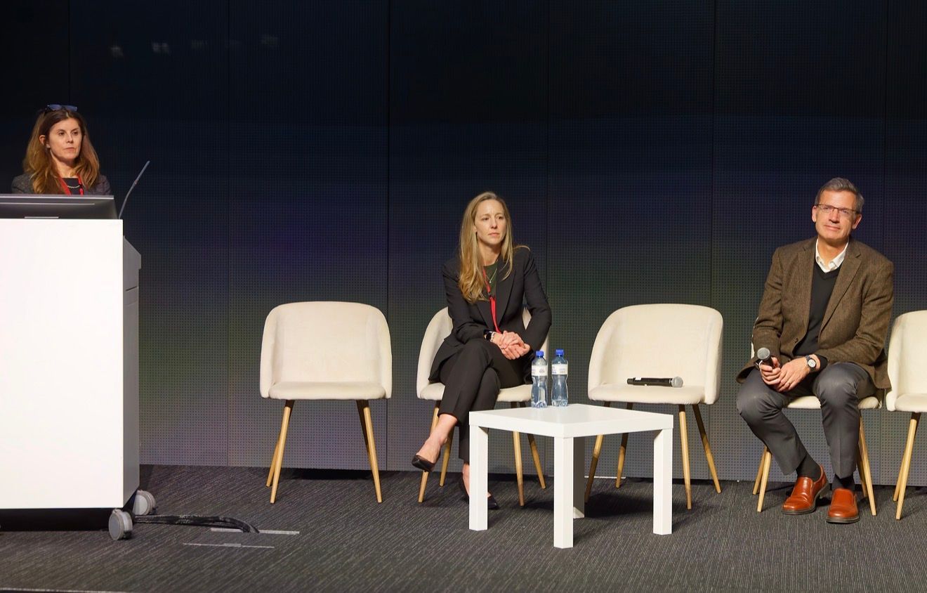 A person stands behind a white podium while two others sit in white chairs on a stage during a panel discussion.