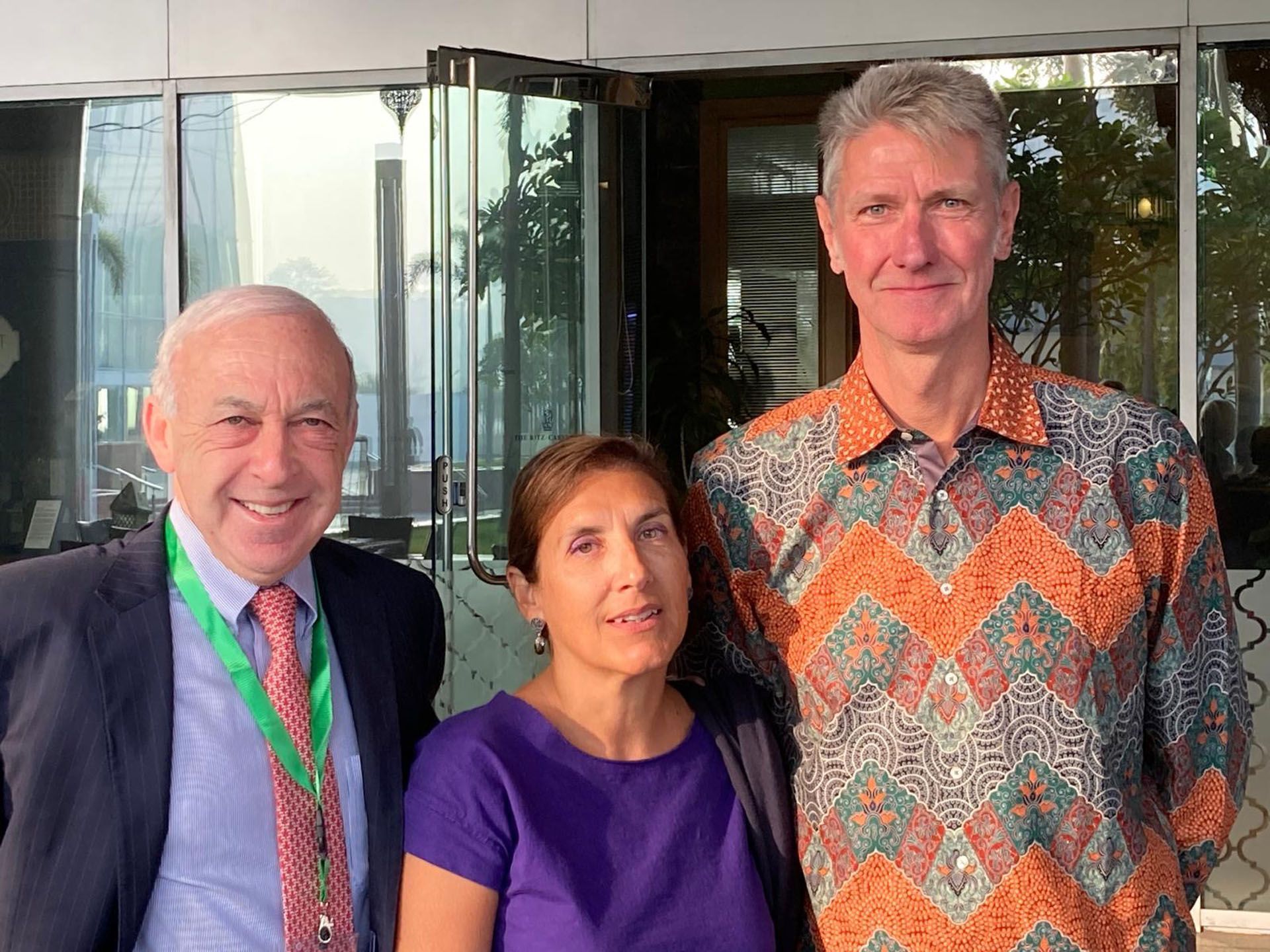 Three people smiling outside a building. Man in suit, woman in purple, man in patterned shirt.