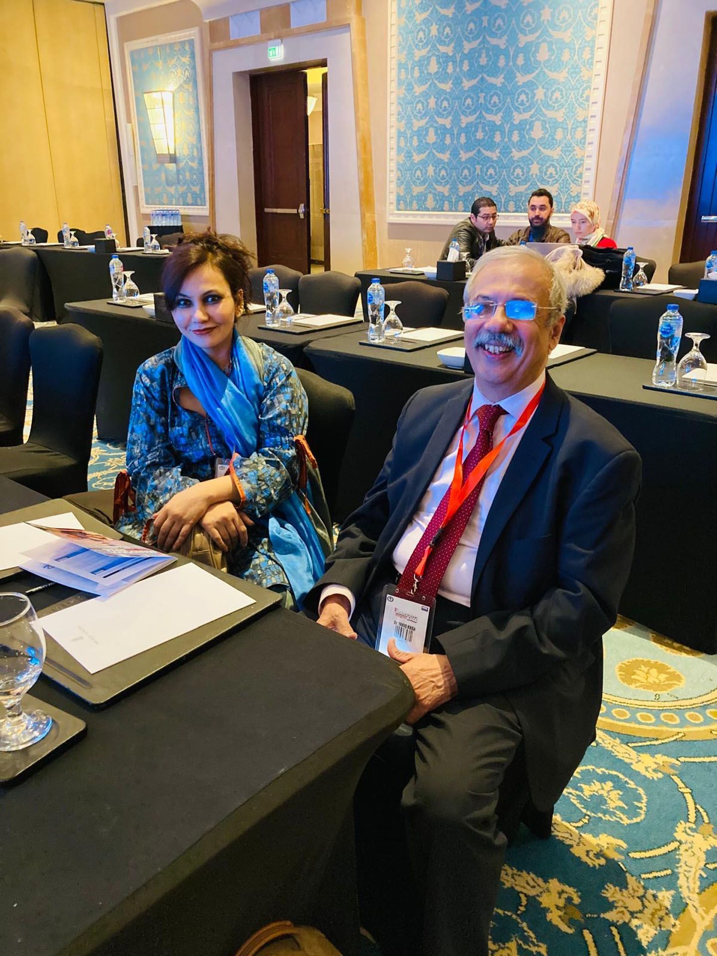 A woman and a man seated at a table in a conference room. Smiling, they look towards the camera.