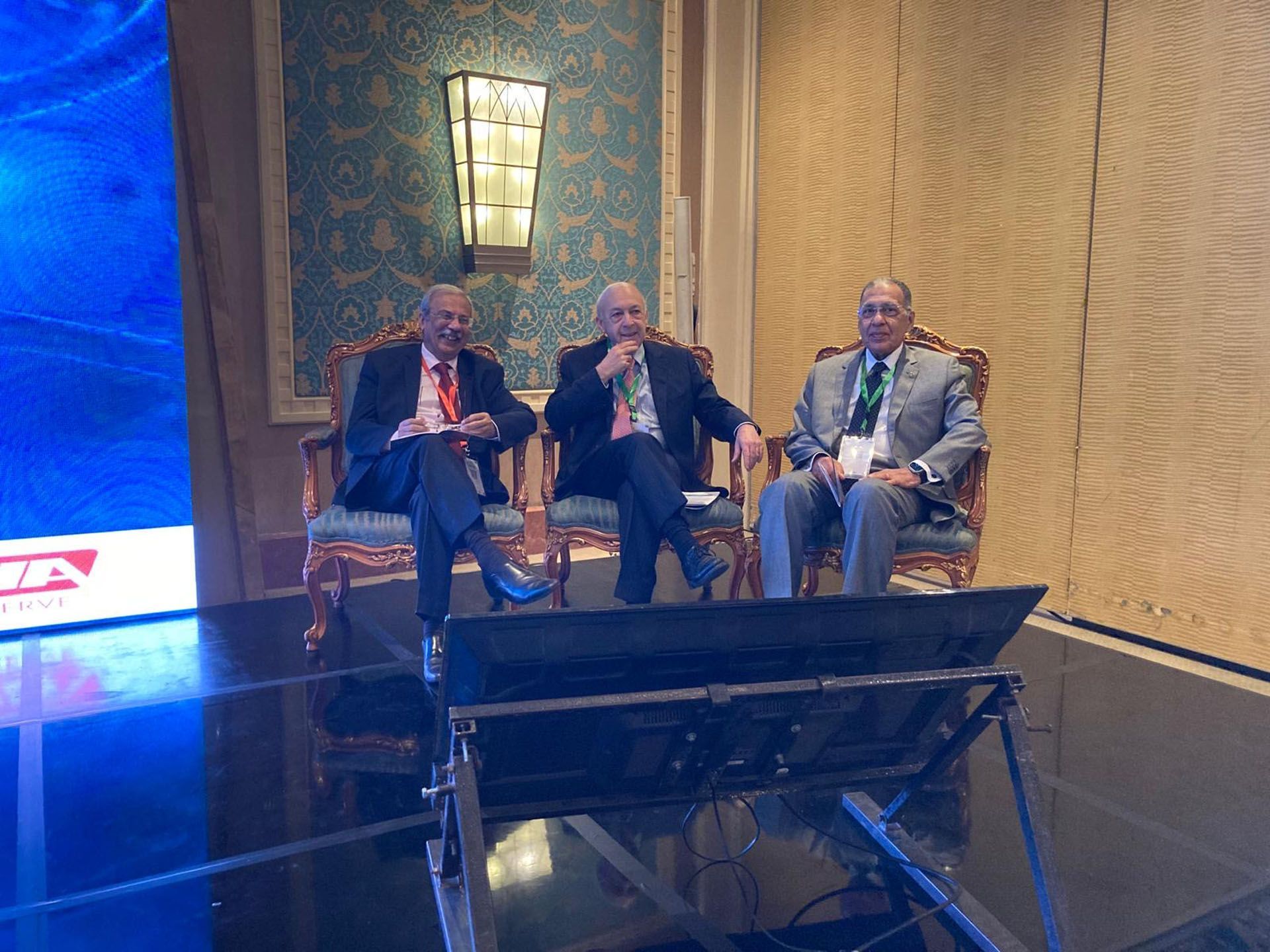 Three men in suits sit on a stage during a conference. They are likely panelists.